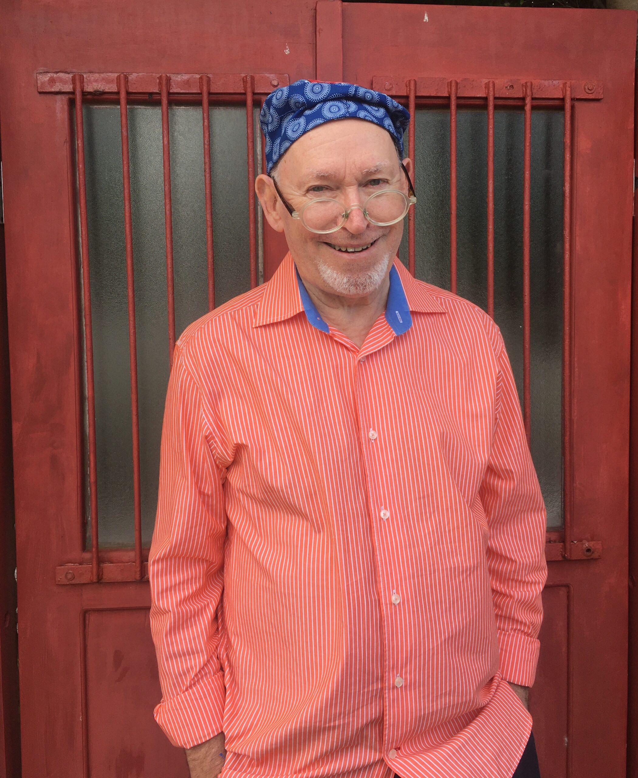 Niki Daly wears a salmon colored shirt and blue bandana standing in front of a wooden door. He smiles at the camera with his glasses perched low on his nose.